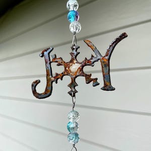 Peut inclure: Un carillon en métal rouillé avec le mot "JOY" et un motif de flocon de neige. Le carillon est orné de perles de verre transparentes et bleues.