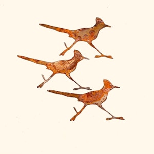 May include: Three rusty metal roadrunner sculptures in a running pose. The sculptures are brown and orange with a rustic finish.
