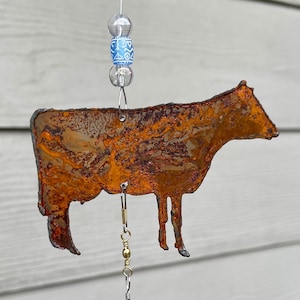 May include: A rusty metal cow wind chime with a blue and white beaded hanger. The cow is brown and has a rustic look.