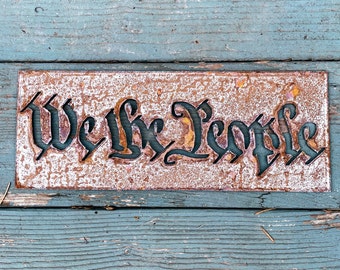 Puede incluir: Un letrero de metal oxidado con las palabras "We the People" recortadas en una fuente decorativa. El letrero está montado en una superficie de madera azul desgastada.