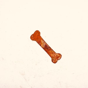 May include: A rusty-orange metal bone-shaped object. The bone has a textured surface with darker brown spots and a rounded shape at each end. The object is set against a plain, off-white background.