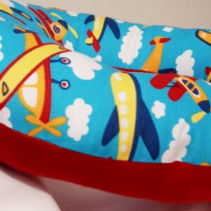 K&ouml;nnte beinhalten: Ein rotes und blaues Donut-f&ouml;rmiges Kissen mit einem leuchtend blauen Hintergrund und einem Muster aus gelben und roten Flugzeugen, wei&szlig;en Wolken und blauem Himmel.