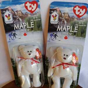 May include: Two white Ty Beanie Baby Maple the Bear plush toys in their original packaging. The packaging features a blue and white background with a picture of a mountain range and a hockey player. The text on the packaging reads "Collect All 4! While supplies last" and "Maple The Bear".
