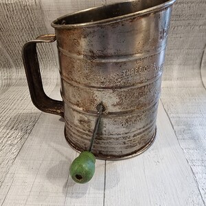 May include: Vintage metal flour sifter with a green wooden handle. The sifter has a handle and a crank to operate it. The sifter is marked with "THREE CUPS MEASURE" and "CUPS 1/2 1 1 1/2 2 2 1/2 3".