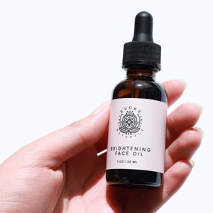 May include: A brown glass bottle with a black dropper top and a white label. The label has a black and white logo with the text "Evoke Naturals" and the text "Brightening Face Oil 1 oz / 30 ml".