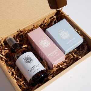 May include: A brown cardboard box filled with shredded paper, containing a brown glass bottle of hydrating body oil, a pink box of brightening face oil, and a blue box of lavender sage soap. The products are labeled with the brand name and product details.
