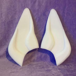 May include: Two white foam elf ears, each with a curved base and a pointed tip. The ears are designed to be worn and are set against a purple background.