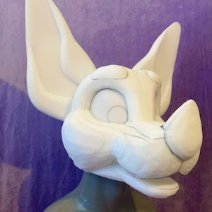 Könnte beinhalten: Ein weißer, unfertiger Fursuit-Kopf mit großen, spitzen Ohren und einer stilisierten Schnauze. Der Kopf ist auf einem Schaufensterpuppenkopf vor einem lila Hintergrund montiert. Das Design enthält sichtbare Umrisse für Details.