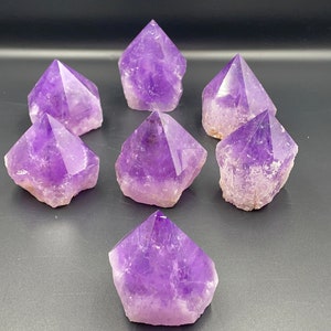 May include: Seven amethyst crystal points in various sizes and shades of purple. The points are arranged in a semi-circle on a dark grey surface.