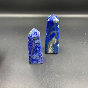May include: Two blue lapis lazuli crystal points, one taller than the other, with white and silver flecks throughout. The points are on a dark grey surface.