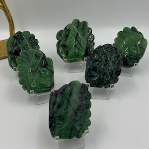 Puede incluir: Seis colgantes de piedra tallada de color verde y rosa que representan la cara de un jefe nativo americano. Los colgantes se muestran en pequeños soportes transparentes.