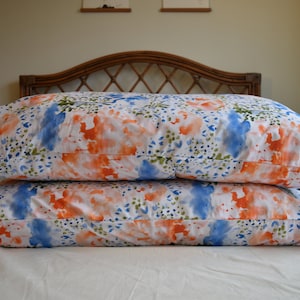 May include: Two white pillowcases with a watercolor pattern of blue, orange, and green. The pillowcases are stacked on top of each other on a white bed.
