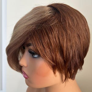May include: A short, brown wig with blonde highlights. The wig is styled in a pixie cut with side-swept bangs.