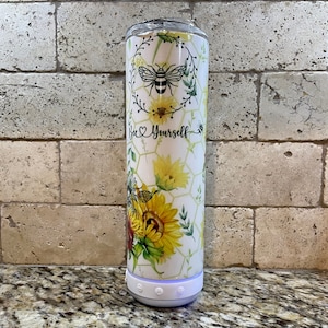 May include: White tumbler with a floral design featuring sunflowers, bees, and a honeycomb pattern. The tumbler has a white lid and a white base with three buttons. The text "Bee Yourself" is printed on the tumbler.