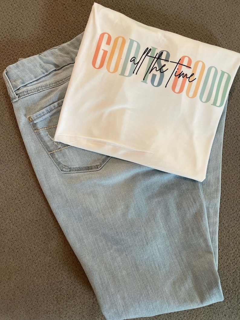 Op de afbeelding: Een wit T-shirt met de tekst "GOD IS GOOD all the time" in regenboogkleuren. Het shirt is gevouwen en ligt op een lichtblauwe spijkerbroek.