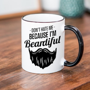 Puede incluir: Taza de cerámica blanca con asa negra y un gráfico negro que dice "Don't hate me - because I'm Beardifull" con una barba negra debajo.
