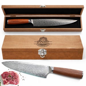 May include: A Damascus steel chef's knife with a wooden handle, presented in a wooden box. The box is engraved with the word "DANIEL" and "PREMIUM QUALITY". The knife is displayed with a piece of raw meat.