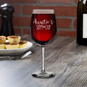 May include: A clear wine glass with a red wine inside. The glass is engraved with the text "Auntie's Sippy Cup" with a small heart symbol.