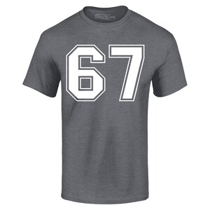 May include: A gray short-sleeve t-shirt with the number "67" in white block letters. The number is outlined in white. The shirt has a crew neck and the brand name "Shop4Ever" is printed on the collar.