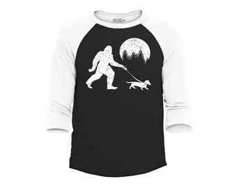 May include: A black and white baseball style raglan shirt with a distressed graphic of Bigfoot walking a dachshund dog on a leash. A full moon and pine trees are in the background. The sleeves are white.