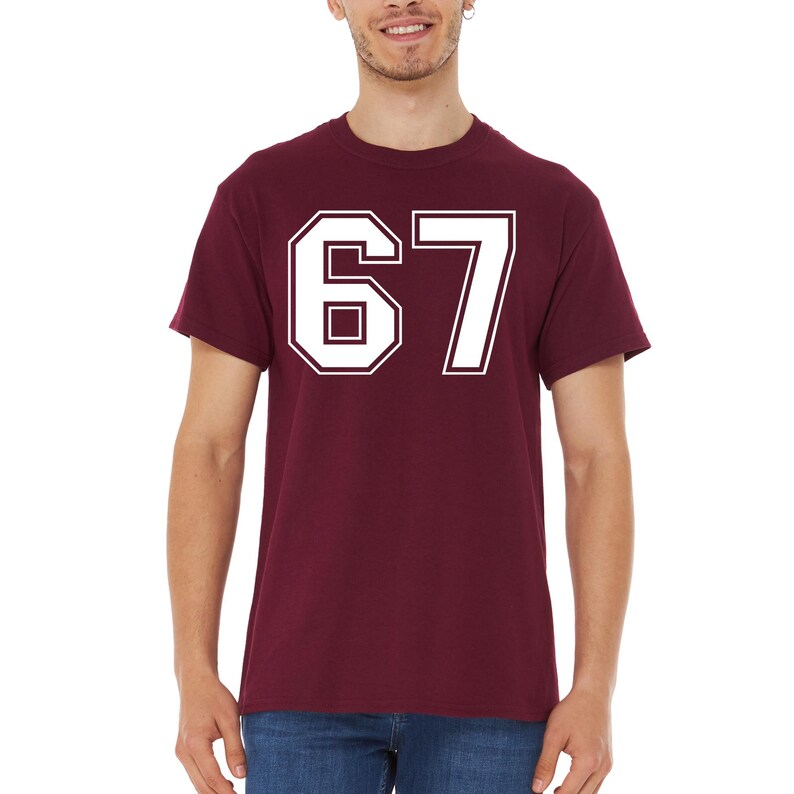 May include: Maroon t-shirt with the number 67 in white, outlined in white. The number is in a collegiate font. The shirt is short-sleeved and made of a soft material.