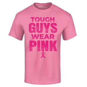 Puede incluir: Camiseta rosa con el texto "Tough Guys Wear Pink" en una fuente rosa desgastada. Una cinta rosa está debajo del texto.