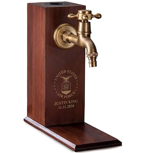May include: A dark brown wooden tap dispenser with a brass faucet. The dispenser has the United States Air Force emblem and the name "JUSTIN KING" with the date "11.11.2026" engraved on the front. The dispenser has a rectangular base.
