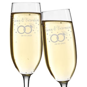 May include: Two champagne flutes filled with a bubbly, golden liquid. Each glass is engraved with the names "Sean & Veronica", interlocking rings, the date "08-02-2025", and small heart and sunburst designs. The glasses are tilted towards each other.