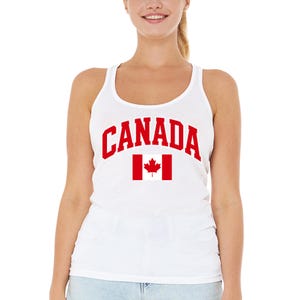 May include: A white tank top featuring the word CANADA printed in bold red capital letters in an arched design above a red and white Canadian flag graphic. The garment has a scoop neckline and wide shoulder straps, providing a simple look.