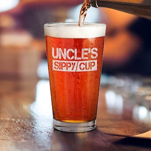 May include: A clear glass pint glass with the text "Uncle's Sippy Cup" etched on the side. The glass is filled with a dark amber colored beer.