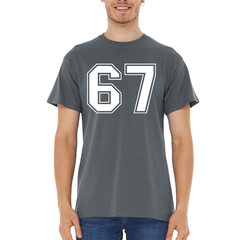 May include: A charcoal gray t-shirt featuring the number "67" in a bold, white, collegiate-style font. The number is outlined with a thick white border. The shirt has short sleeves and a crew neck.
