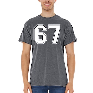 May include: A charcoal gray t-shirt featuring the number 67 in a white, bold, block font. The shirt has short sleeves and a crew neck. The number is centered on the chest.