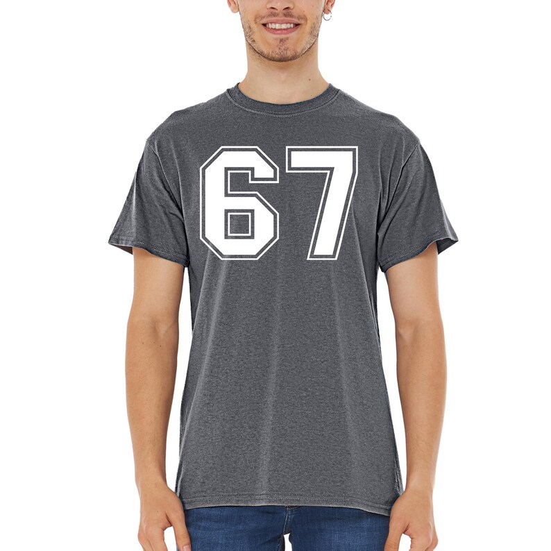 May include: A charcoal gray t-shirt featuring the number 67 in a white, bold, block font. The shirt has short sleeves and a crew neck. The number is centered on the chest.