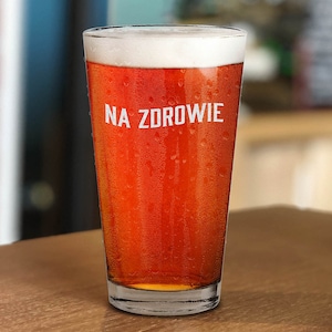 Puede incluir: Un vaso de pinta de vidrio transparente lleno de una cerveza marrón rojiza. El vaso tiene condensación en el exterior y el texto "NA ZDROWIE" impreso en el lateral.