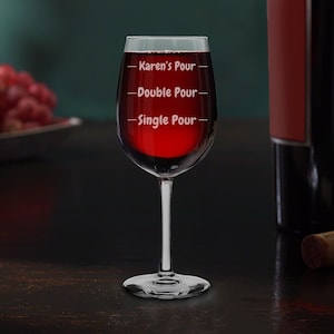 May include: A clear glass wine glass with a red wine pour. The glass has three lines etched on the side with the text "Karen's Pour", "Double Pour", and "Single Pour".