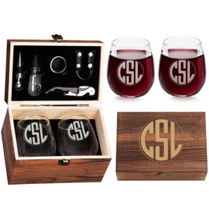 Op de afbeelding: Een houten wijngeschenkset met twee wijnglazen zonder voet met de initialen "CSL" gegraveerd, een houten doos en wijnaccessoires. De doos en de glazen zijn gevuld met rode wijn.