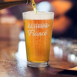 May include: A clear pint glass filled with golden beer and white foam. The glass has the words "Boyfriend Fiance" etched in a cursive font. Beer is being poured into the glass from a metal spout.
