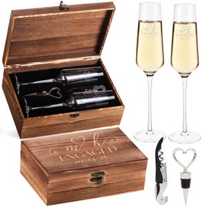 May include: A wooden gift box set with a champagne theme. The box is engraved with "m k ENGAGED 02-14-26". Inside are two champagne flutes, a bottle stopper with a heart design, and a corkscrew. Two filled champagne flutes are also shown.