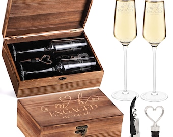 May include: A wooden gift box set with a champagne theme. The box is engraved with "m k ENGAGED 02-14-26". Inside are two champagne flutes, a bottle stopper with a heart design, and a corkscrew. Two filled champagne flutes are also shown.