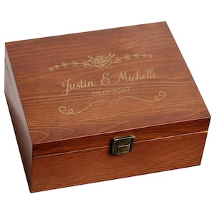 May include: A wooden box with a hinged lid and a metal clasp. The box is engraved with the names "Justin & Michelle" and the date "09.01.2030", along with floral and scroll designs. The wood has a warm, reddish-brown tone.