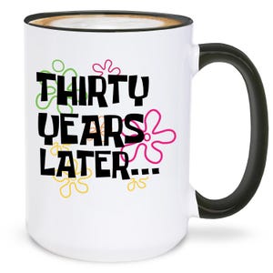 May include: White ceramic mug with a black handle and rim, featuring the text "THIRTY YEARS LATER..." in black, with colorful flower accents. The mug contains a coffee-like beverage.
