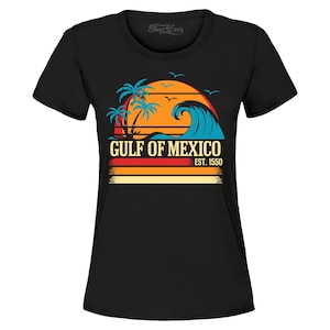 Puede incluir: Camiseta negra con un diseño gráfico colorido de una puesta de sol sobre el Golfo de México. El diseño incluye palmeras, una ola y el texto "GULF OF MEXICO EST. 1550". La camiseta tiene mangas cortas y cuello redondo.