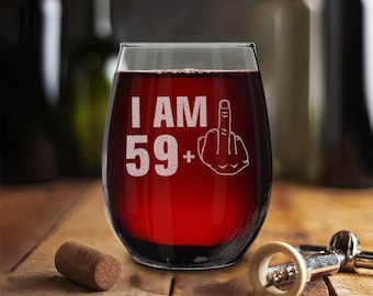 Op de afbeelding: Een wijnglas zonder steel met de tekst "I AM 59+" en een middelvinger-emoji gegraveerd aan de zijkant. Het glas is gevuld met rode wijn.