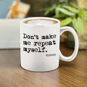 May include: White ceramic mug with a handle, filled with a coffee beverage and foam. The mug features the text "Don't make me repeat myself." below which is the word "-History" in a smaller font. The mug is on a wooden surface.