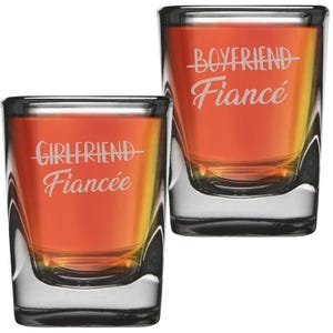 May include: Two clear shot glasses filled with an orange liquid. One glass has the words "Girlfriend" and "Fiancée" etched on it, while the other says "Boyfriend" and "Fiancé". The glasses have a square base and a slightly flared top.