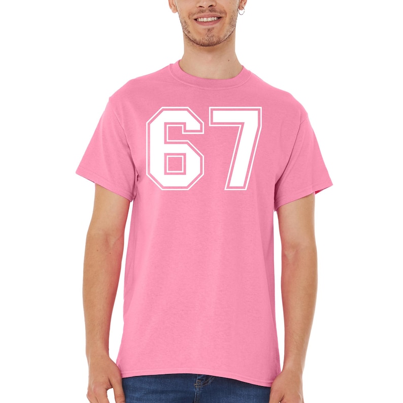 May include: A pink t-shirt with the number 67 in white, featuring a bold, outlined font. The shirt is a solid color, with a crew neck and short sleeves. The number is centered on the chest.