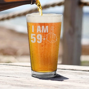 May include: A clear glass pint glass with the text "I AM 59+" and a middle finger emoji etched on the side. The glass is filled with a light amber colored beer.