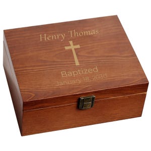 May include: A wooden box with a hinged lid and a metal clasp. The box is stained a warm brown color and features the engraved text "Henry Thomas", a cross, and "Baptized January 18, 2030". The box is likely used for keepsakes or gifts.