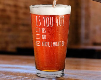 Op de afbeelding: Een helder glazen pintglas met de tekst "IS YOU 40?" en een checklist met de opties "YES", "NO" en "BITCH, I MIGHT BE". Het glas is gevuld met een roodbruine vloeistof.