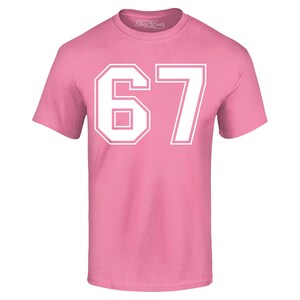 May include: A pink t-shirt with the number "67" in white block letters. The number is outlined in white and centered on the front of the shirt. The shirt has short sleeves and a crew neck.
